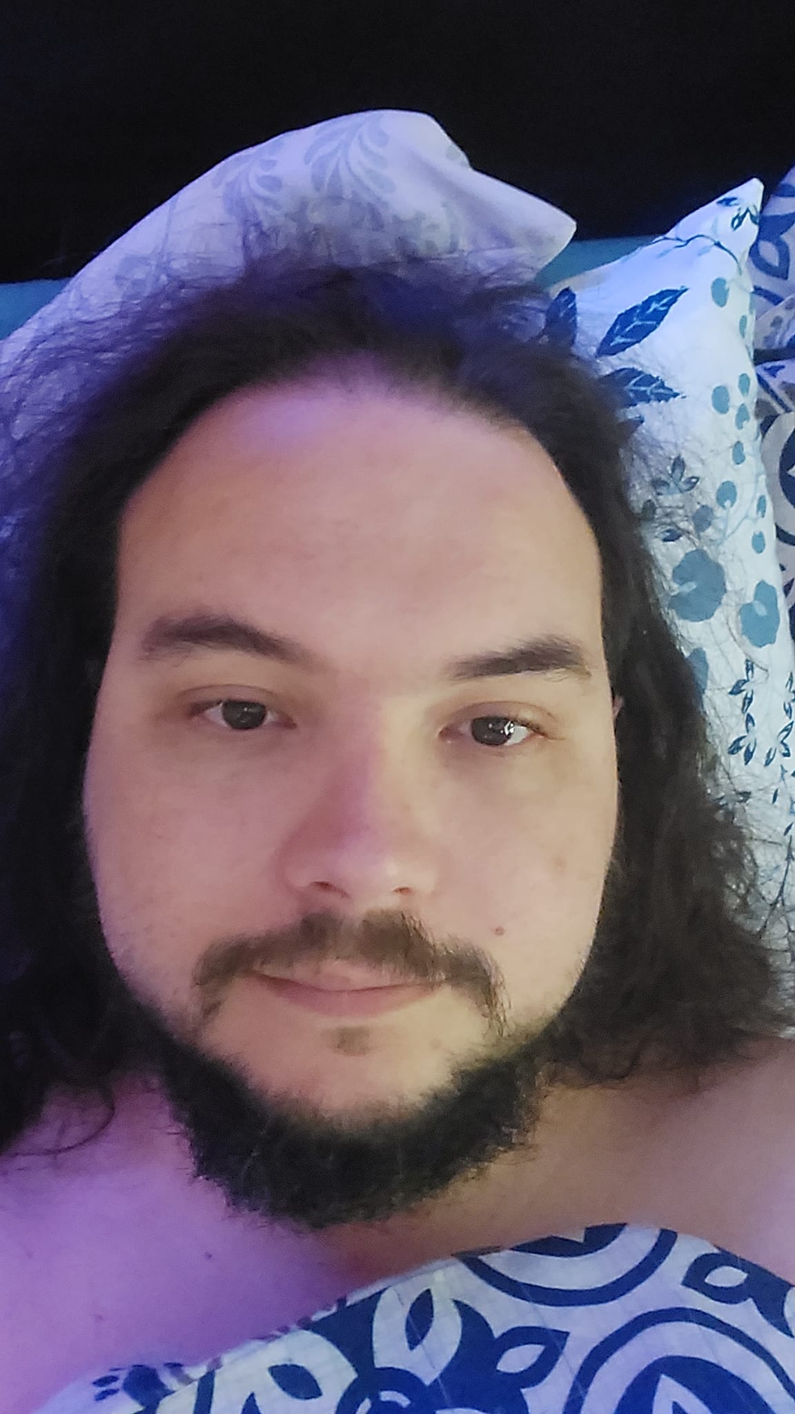 A man with long dark hair and a beard lies in bed